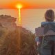 A person with a backpack and a small dog stand on a coastal trail at sunset, overlooking the ocean with the sun low on the horizon.