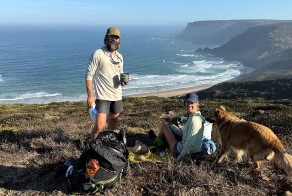 Two people and a dog sit on a grassy cliff overlooking the ocean, with backpacks and supplies beside them on a sunny day.