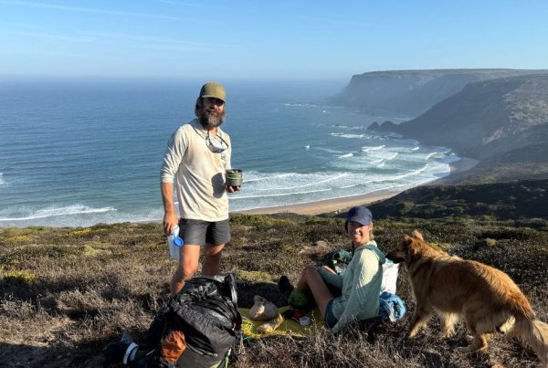 Two people and a dog sit on a grassy cliff overlooking the ocean, with backpacks and supplies beside them on a sunny day.
