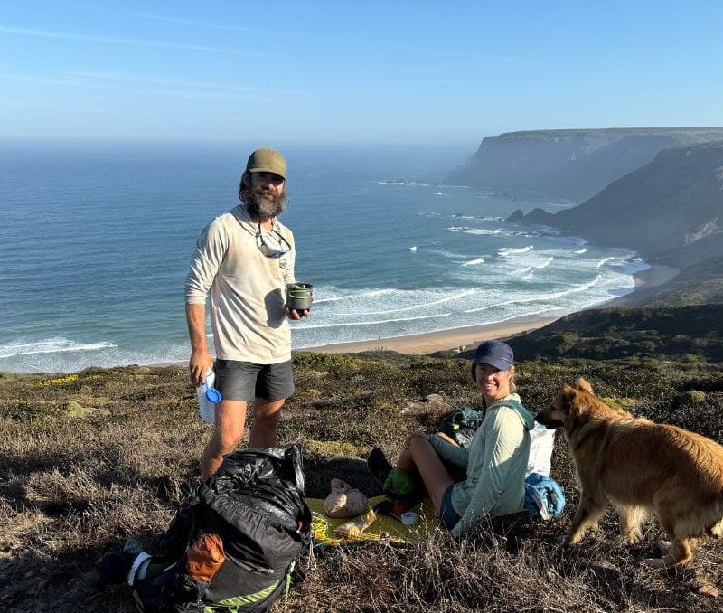 Two people and a dog sit on a grassy cliff overlooking the ocean, with backpacks and supplies beside them on a sunny day.