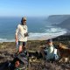 Two people and a dog sit on a grassy cliff overlooking the ocean, with backpacks and supplies beside them on a sunny day.