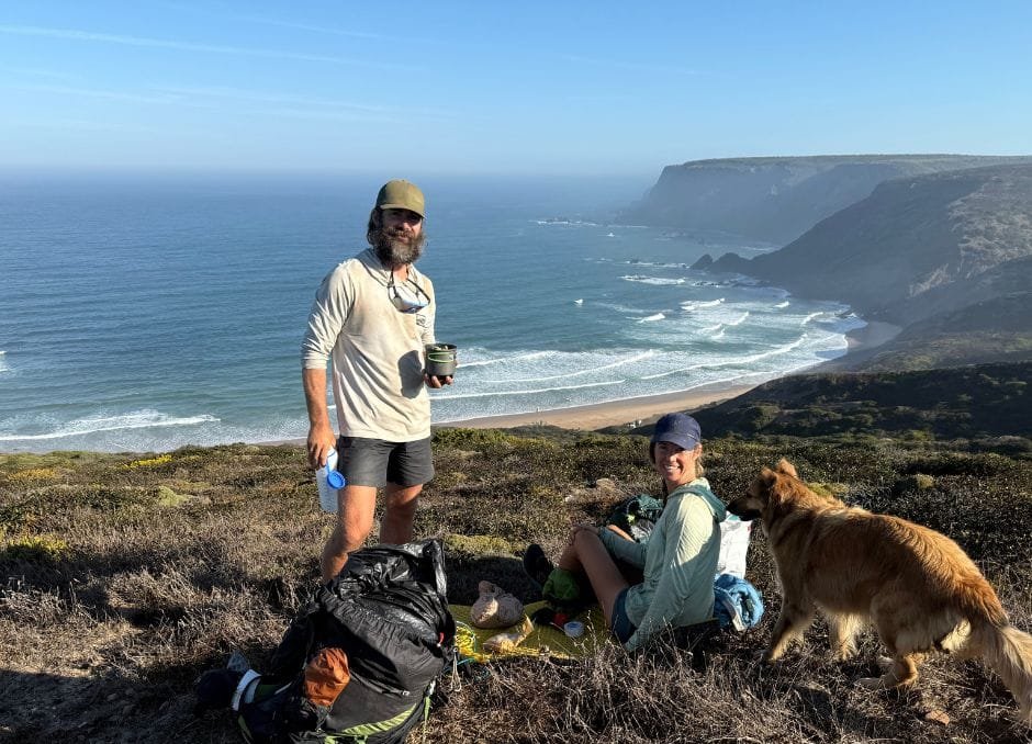 Two people and a dog sit on a grassy cliff overlooking the ocean, with backpacks and supplies beside them on a sunny day.