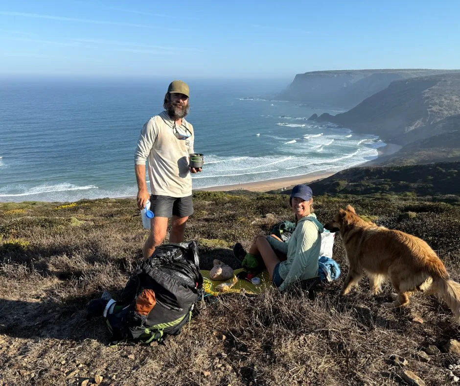 Two hikers and a dog resting on a grassy cliff overlooking the Atlantic on the Fisherman's Trail.