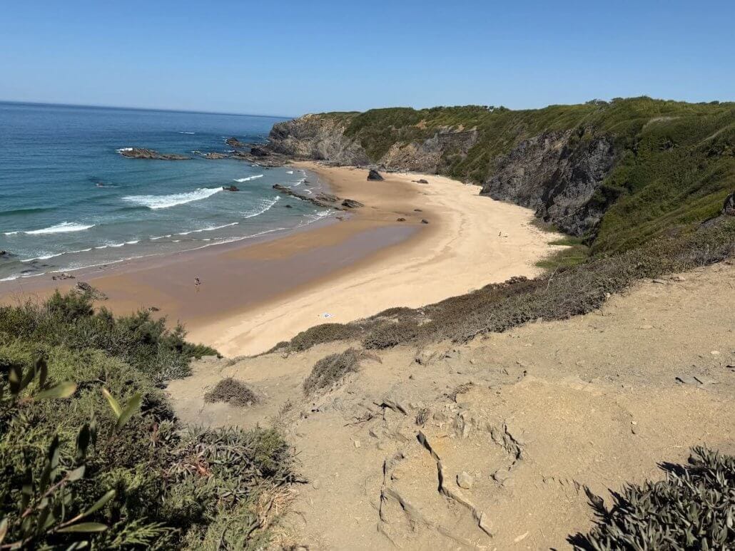A sandy beach bordered by rocky cliffs and green vegetation, with gentle waves and a few people walking along the shoreline under a clear blue sky.