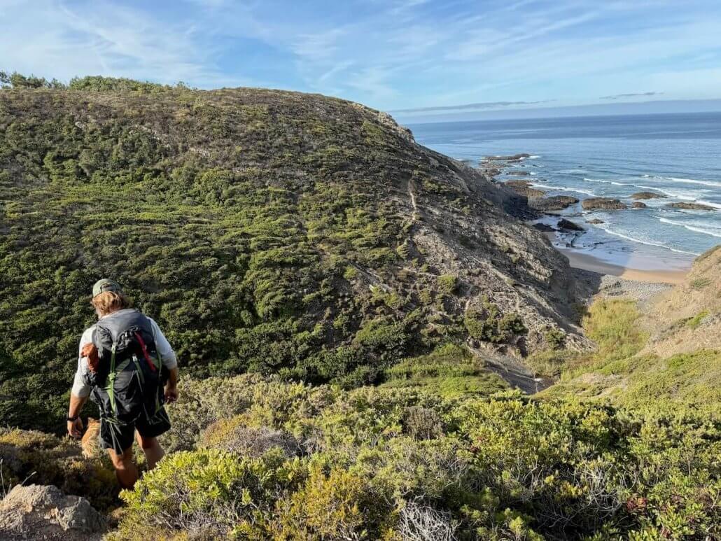 Two hikers with backpacks walk down a grassy, rocky hill toward a coastal beach with waves and cliffs under a blue sky.