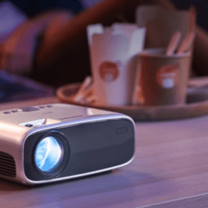 A small, portable projector is turned on and placed on a table, with takeout food containers and people sitting in the background.
