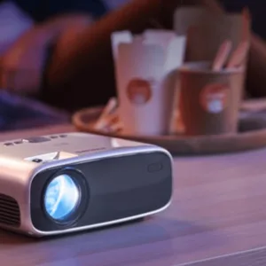A small, portable projector is turned on and placed on a table, with takeout food containers and people sitting in the background.