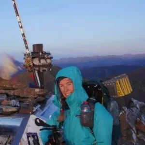 Wearing the Rain / Wind Jacket, a person stands at a mountain summit next to a cross and gear, smiling with breathtaking mountains in the background.