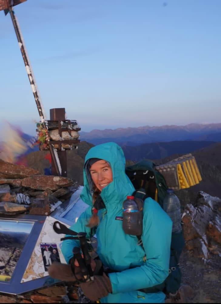 Wearing the Rain / Wind Jacket, a person stands at a mountain summit next to a cross and gear, smiling with breathtaking mountains in the background.