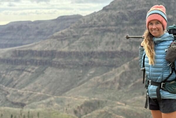 A woman wearing hiking gear and a pink hat stands with trekking poles on her shoulder, smiling, with rocky mountains in the background.