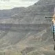 A woman wearing hiking gear and a pink hat stands with trekking poles on her shoulder, smiling, with rocky mountains in the background.
