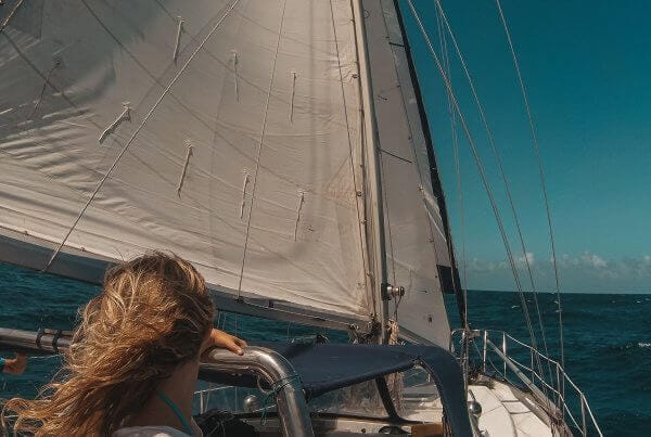 A person with long hair, seen from behind, steering a sailboat on the open ocean under clear blue skies.