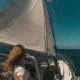 A person with long hair, seen from behind, steering a sailboat on the open ocean under clear blue skies.