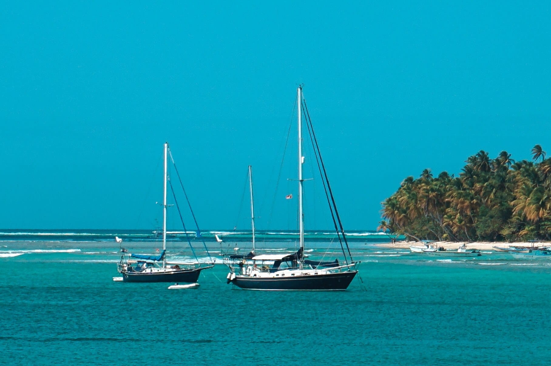 Two sailboats are anchored in clear blue water near a tropical island with palm trees under a bright blue sky.