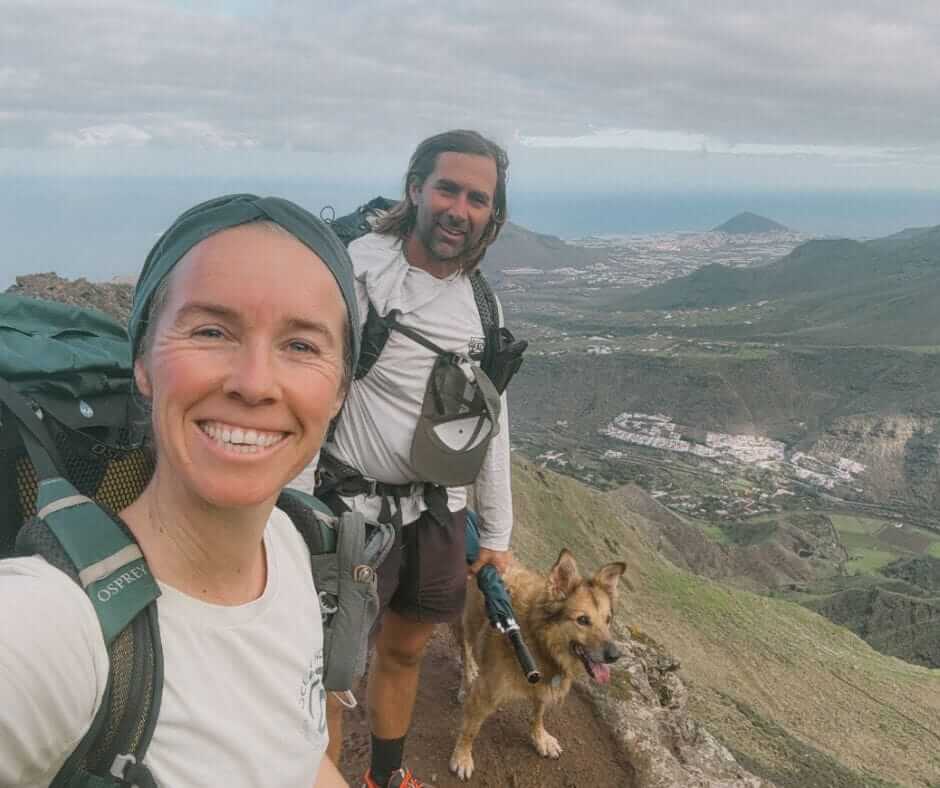 Two people with backpacks and a dog stand on a mountain trail with a green valley, towns, and the ocean visible in the background.