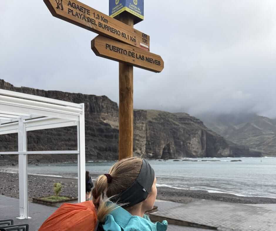 A person with a backpack stands by a wooden signpost near a rocky beach, with cliffs and cloudy skies in the background. The sign points to nearby locations.