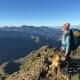 A hiker with a backpack and a dog stands on a rocky mountain ridge, overlooking a vast landscape of mountains under a clear blue sky.