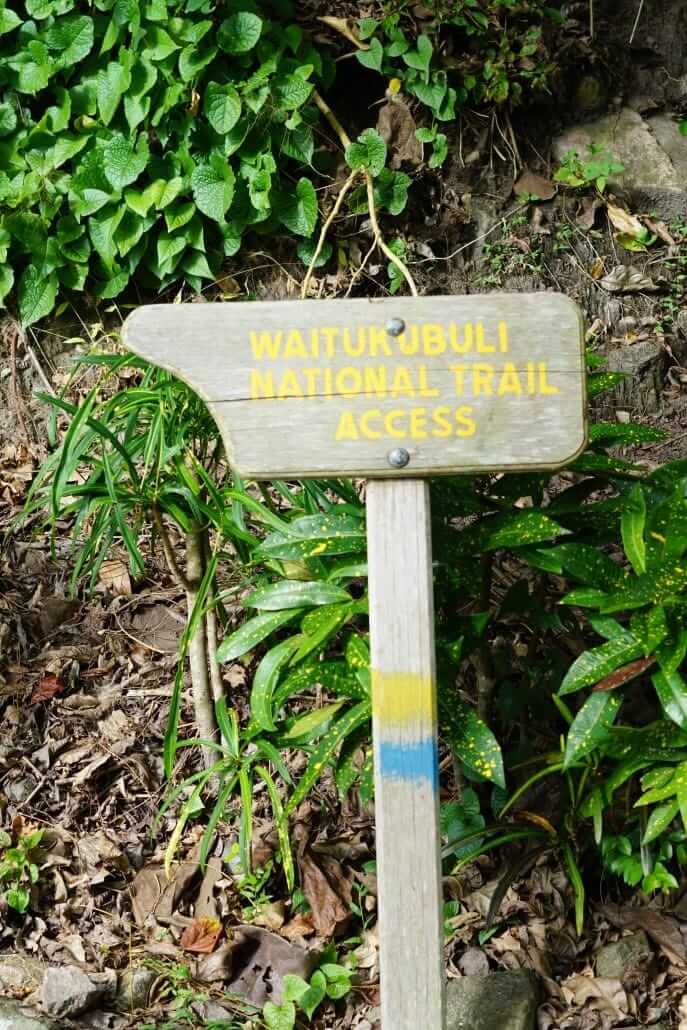 A wooden sign reads "Waitukubuli National Trail Access" and is surrounded by green foliage and leaves on the ground.