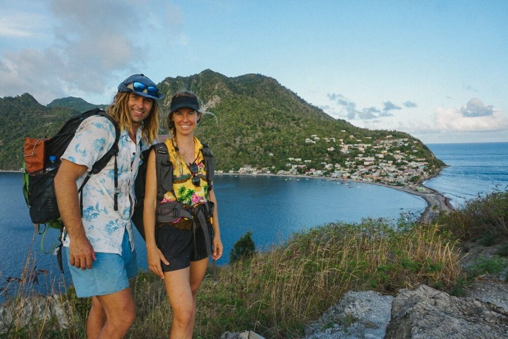 Two hikers wearing backpacks and casual clothes stand on a hill overlooking a coastal village, with the sea and a green, mountainous landscape in the background.