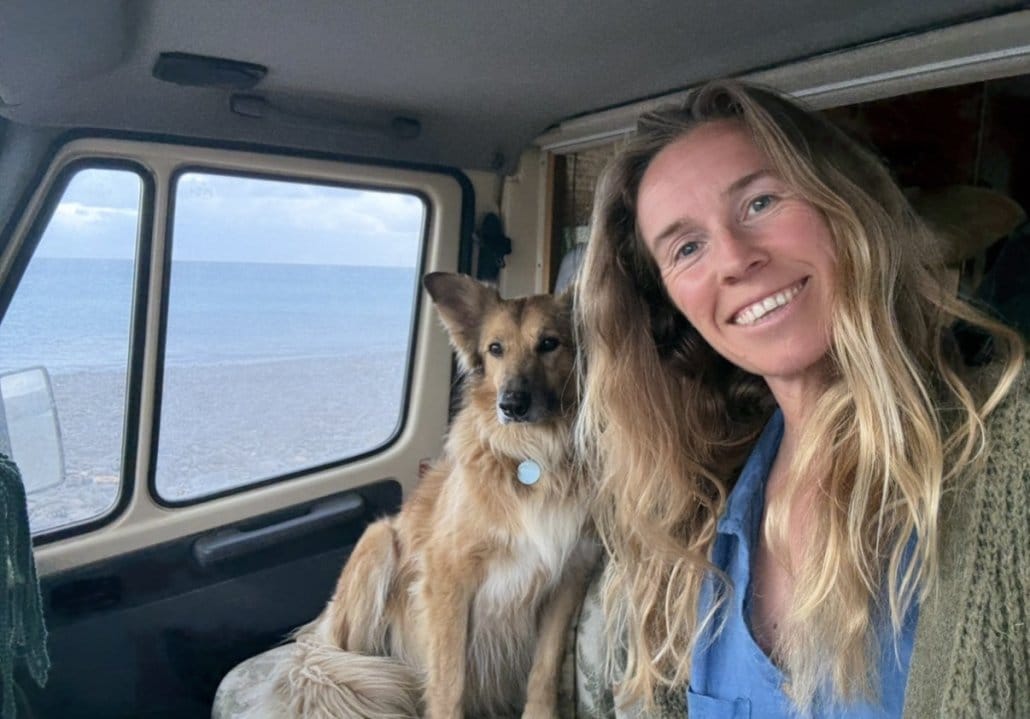 A woman and a dog sit inside a vehicle parked by a beach, with the ocean visible through the window.