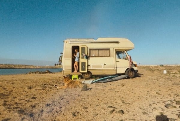 A woman stands in the doorway of a beige camper van parked on a sandy, rocky beach with a dog lying nearby and surfboards on the ground.