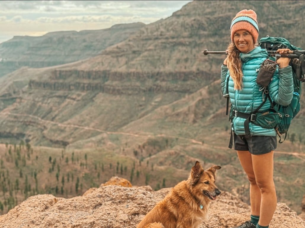 Woman in hiking gear stands on a rocky ledge with a backpack and trekking poles, smiling; a brown dog sits beside her with a mountainous landscape in the background.