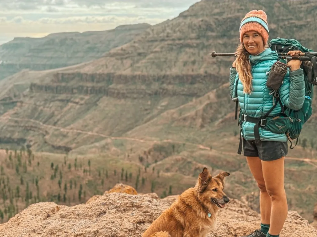 Woman in hiking gear stands on a rocky ledge with a backpack and trekking poles, smiling; a brown dog sits beside her with a mountainous landscape in the background.