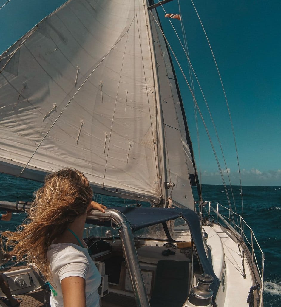Person with long hair holds onto railing while sailing on a boat under clear blue sky, with large sail and ocean visible.