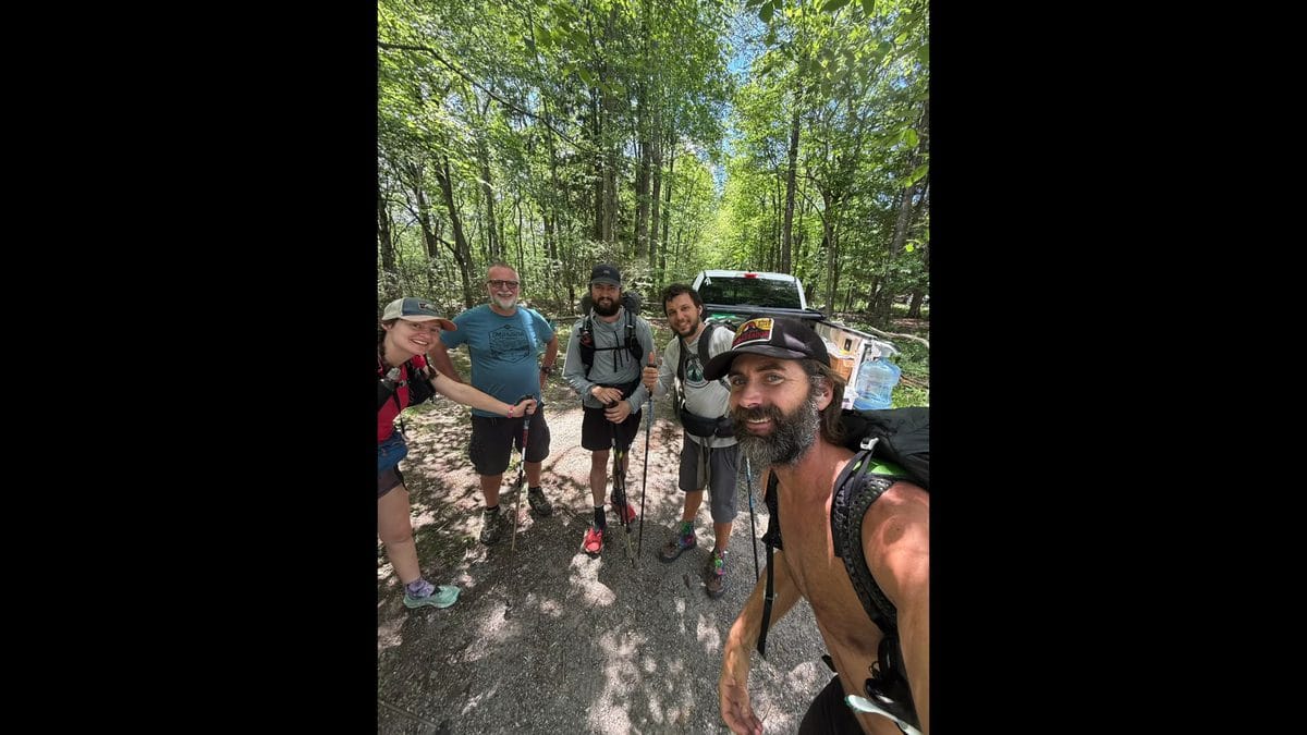 A group of six hikers posing together on the Appalachian Trail, wearing backpacks and trail gear, smiling