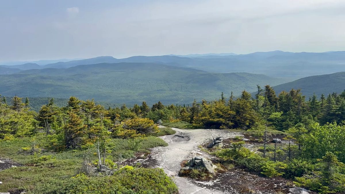 High-elevation rocky Appalachian Trail with low vegetation and distant layered mountain ranges under a clear blue sky