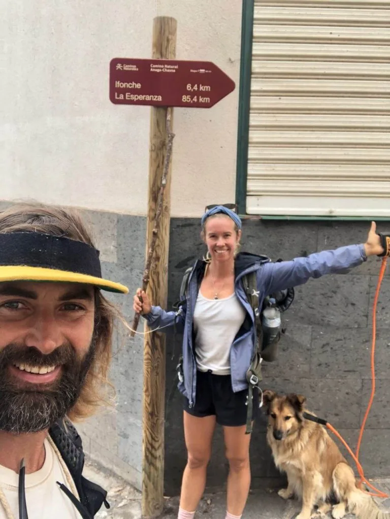 Two hikers with backpacks stand next to a signpost showing distances to Ifonche and La Esperanza; a dog on an orange leash sits beside them.