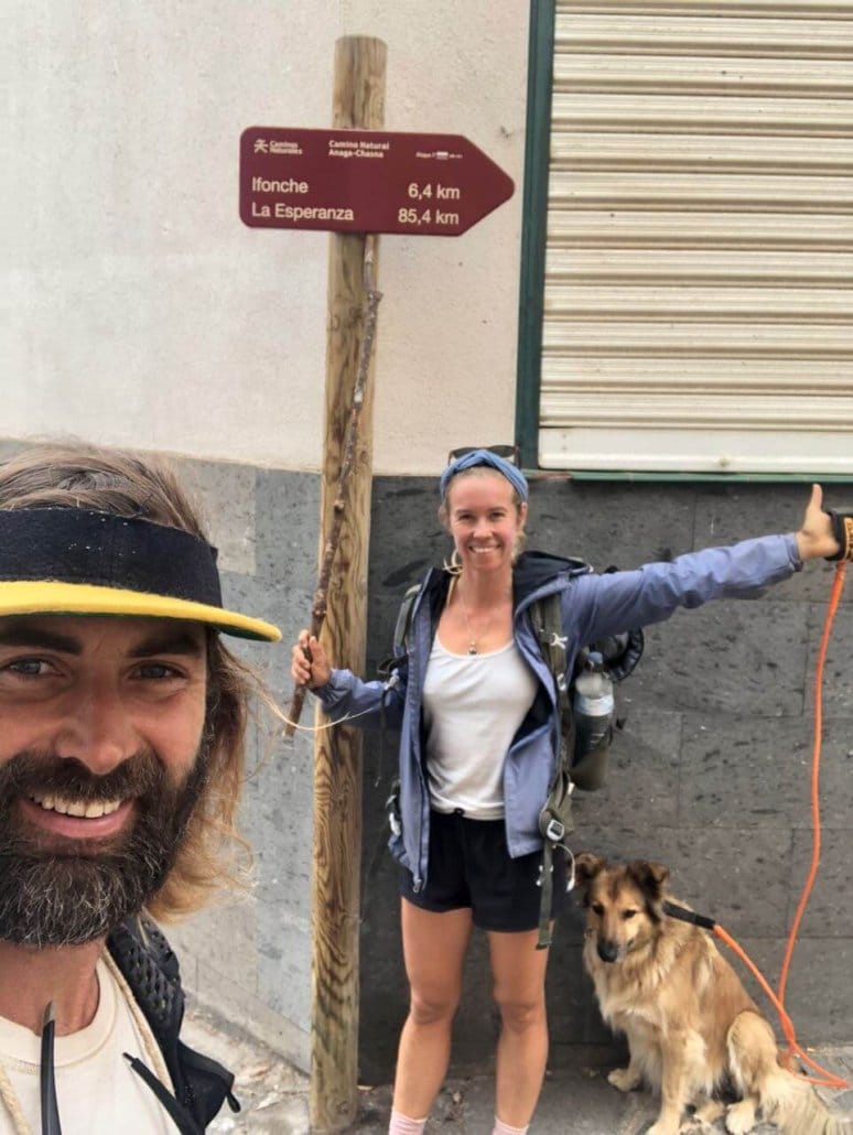 Two hikers with backpacks stand next to a signpost showing distances to Ifonche and La Esperanza; a dog on an orange leash sits beside them.