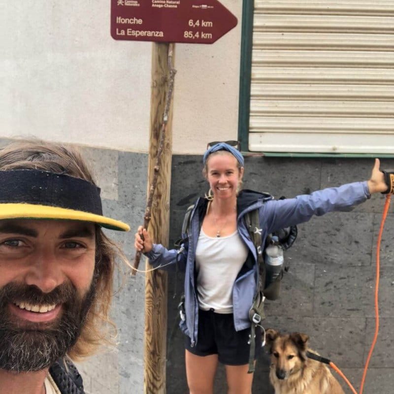 Two hikers with backpacks stand next to a signpost showing distances to Ifonche and La Esperanza; a dog on an orange leash sits beside them.