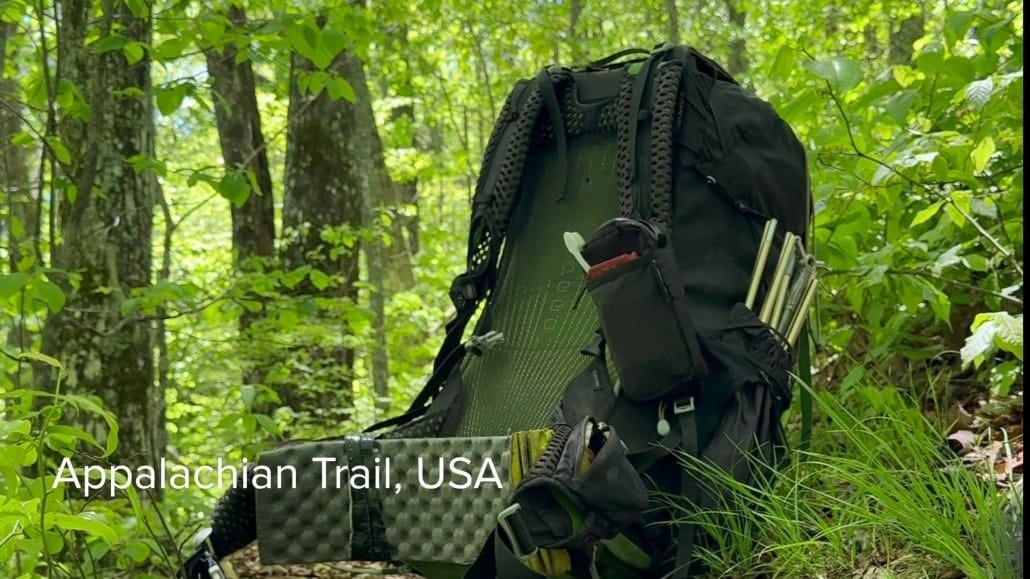 A black hiking backpack with gear sits on the forest floor surrounded by green foliage. Text reads: "Appalachian Trail, USA.