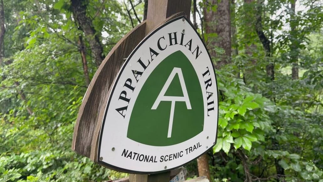 A wooden sign with a green and white Appalachian Trail logo in a forested area, labeled "National Scenic Trail.