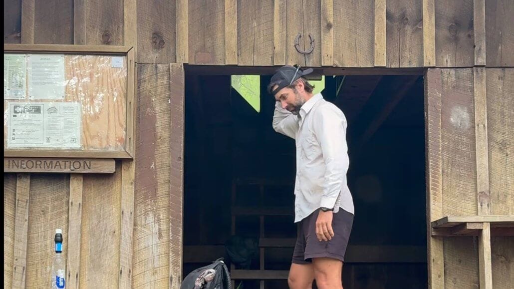 A person in outdoor clothing stands in the doorway of a wooden cabin, adjusting their hat. A backpack and information board are visible nearby.