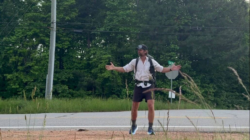 A person wearing a cap, backpack, and athletic clothes stands with arms outstretched near a roadside, with trees and power lines in the background.