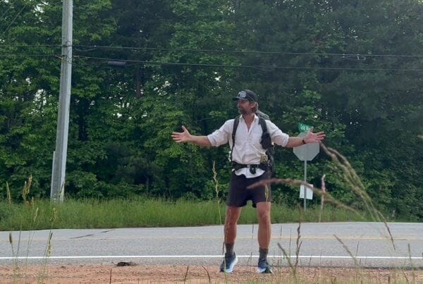 A person wearing a cap, backpack, and athletic clothes stands with arms outstretched near a roadside, with trees and power lines in the background.