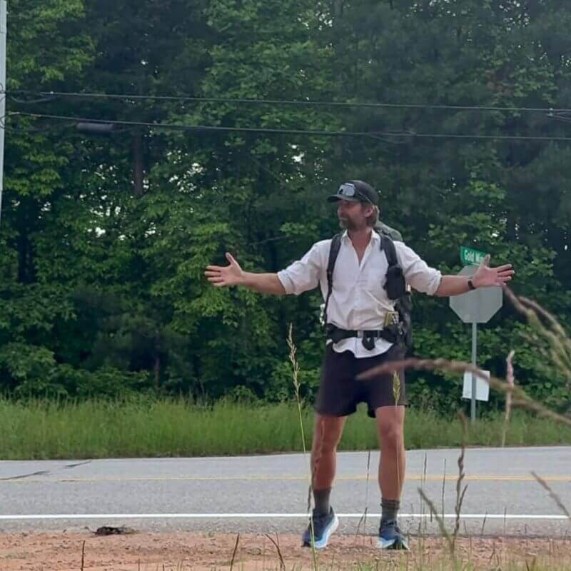 A person wearing a cap, backpack, and athletic clothes stands with arms outstretched near a roadside, with trees and power lines in the background.