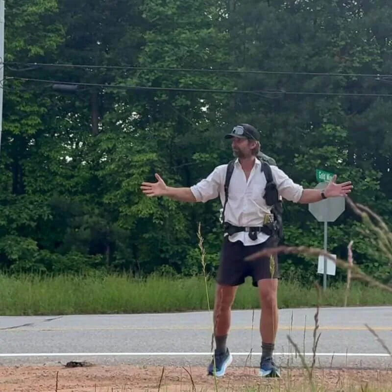 A person wearing a cap, backpack, and athletic clothes stands with arms outstretched near a roadside, with trees and power lines in the background.