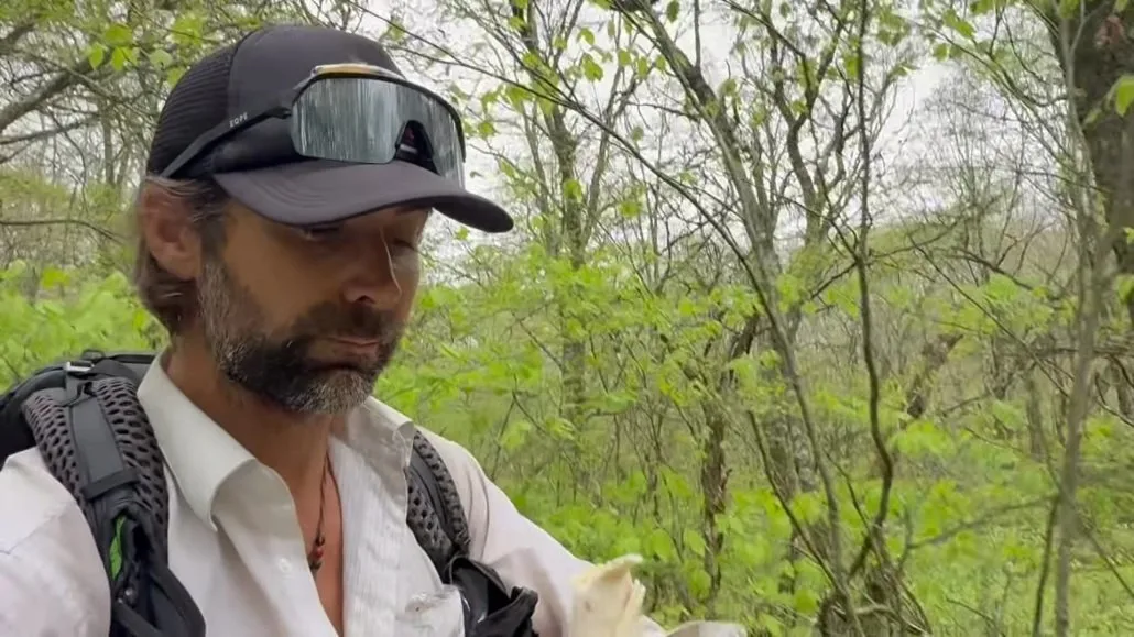 A man wearing a black cap, sunglasses, and a white shirt stands in a green, wooded area, holding a sandwich.