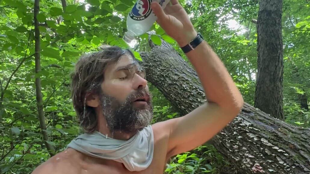 A man with a gray scarf around his neck pours water from a plastic bottle over his head in a forested area, cooling off in the shade.