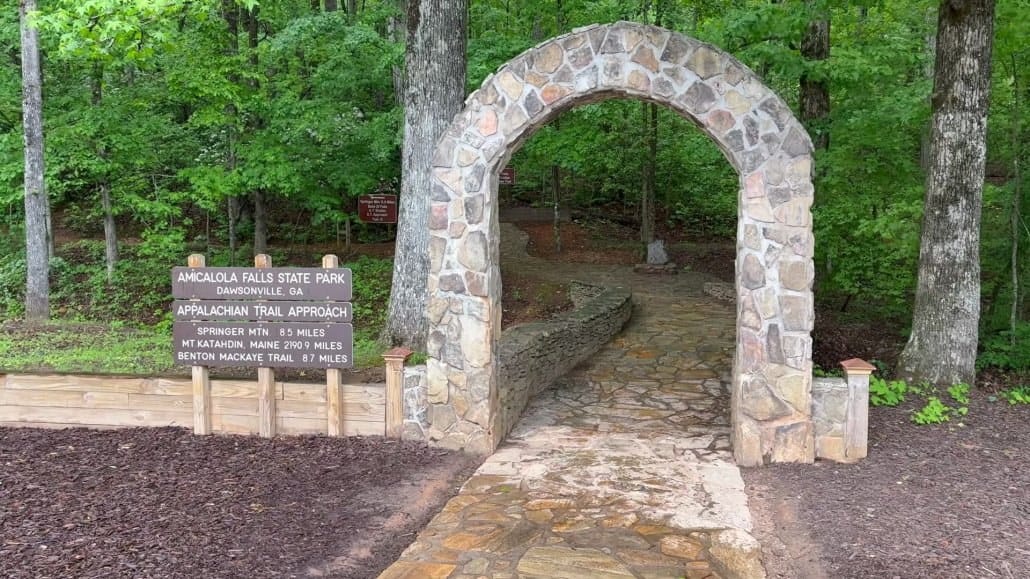 Stone archway entrance to a forest trail at Amicalola Falls State Park, with signs indicating the start of the Appalachian Trail Approach and distances to various trail points.