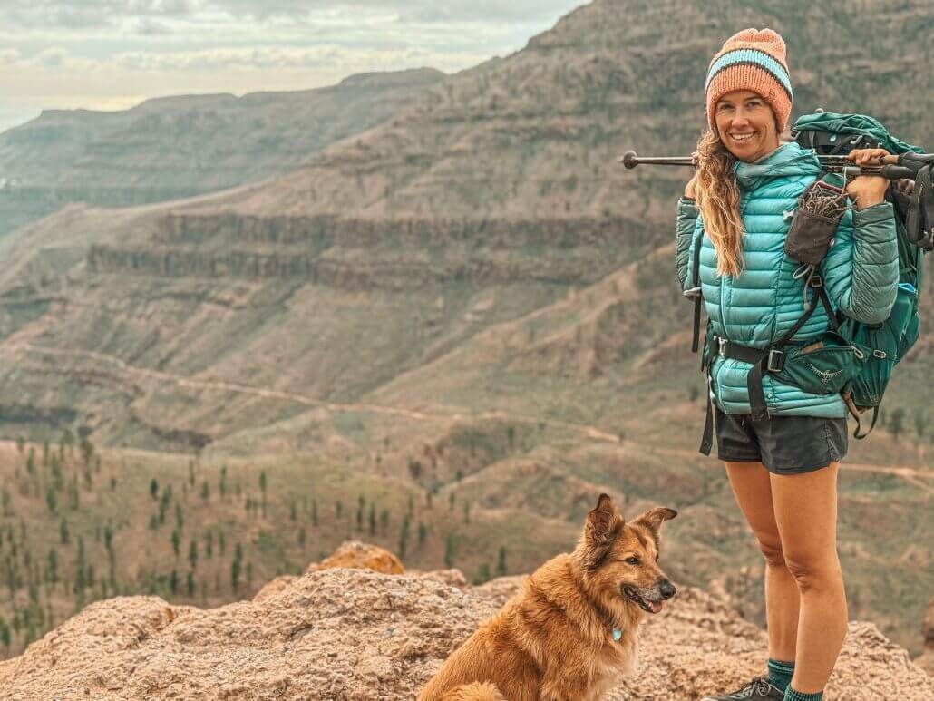 A woman in a teal jacket and beanie stands on a rocky cliff with hiking gear, next to a brown dog, overlooking a vast canyon landscape.