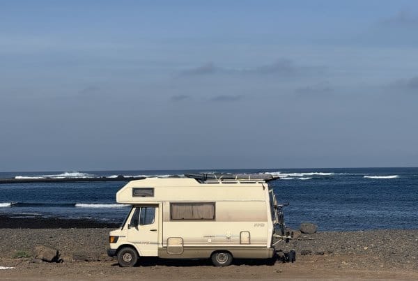 Vintage Mercedes camper with a surfboard on the roof parked on a rocky Fuerteventura shore, waves breaking behind.