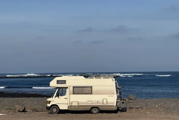Vintage Mercedes camper with a surfboard on the roof parked on a rocky Fuerteventura shore, waves breaking behind.