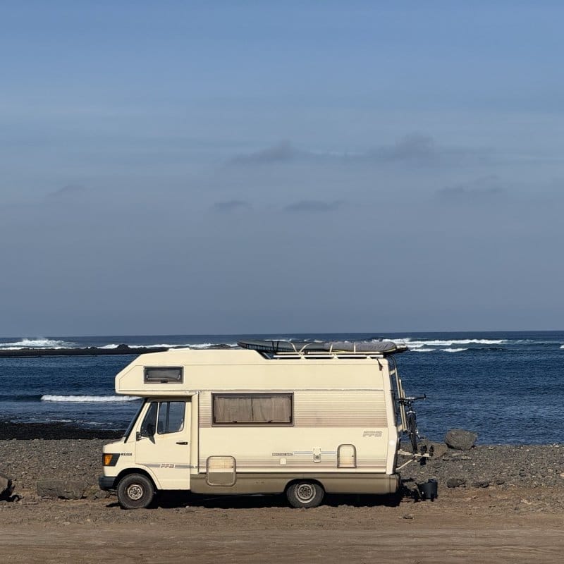 Vintage Mercedes camper with a surfboard on the roof parked on a rocky Fuerteventura shore, waves breaking behind.