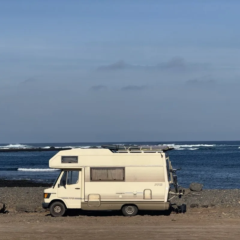 Vintage Mercedes camper with a surfboard on the roof parked on a rocky Fuerteventura shore, waves breaking behind.
