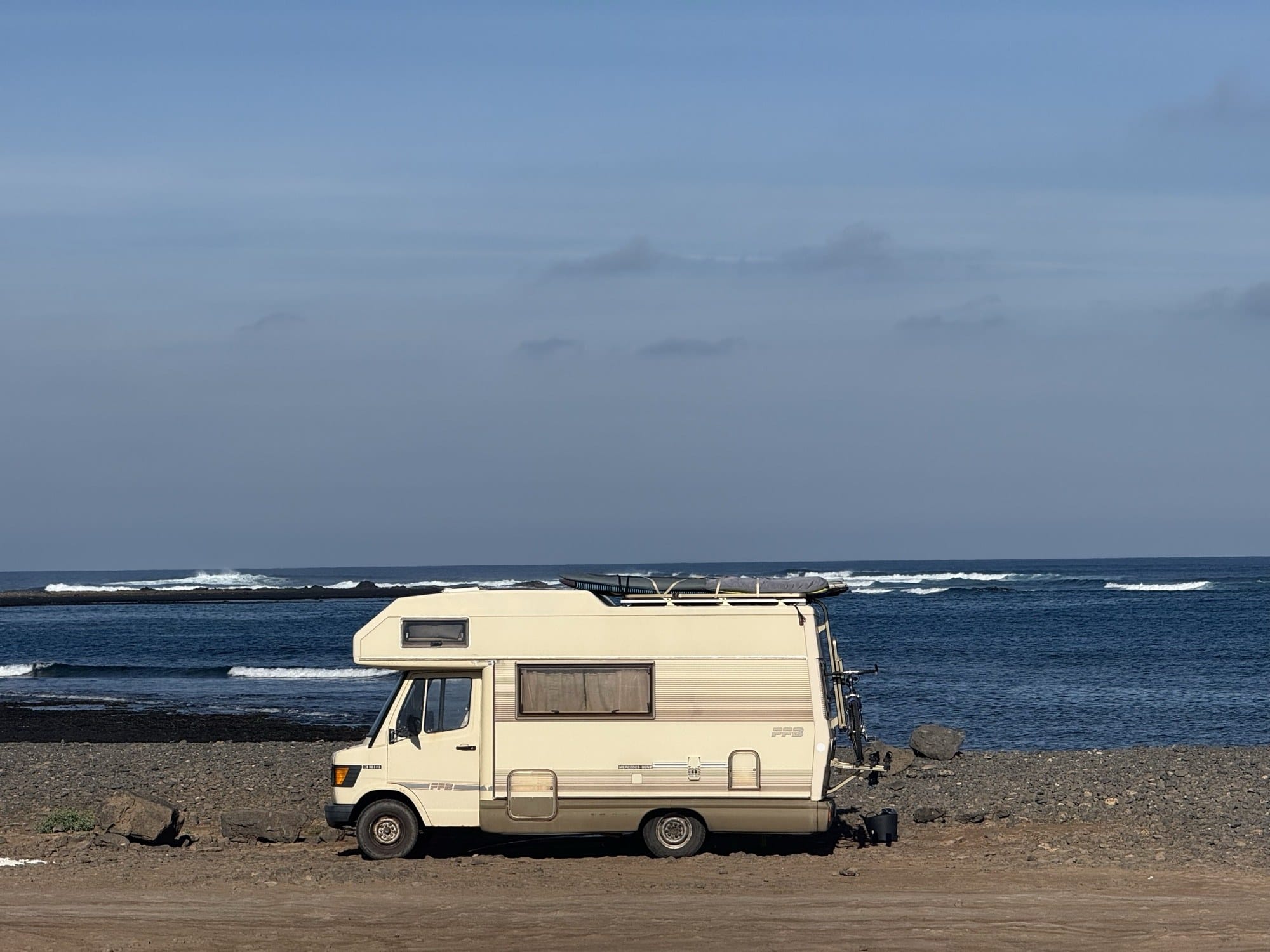 Vintage Mercedes camper with a surfboard on the roof parked on a rocky Fuerteventura shore, waves breaking behind.